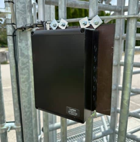 Työmaan työntekijä keltaisessa heijastintakissa työmaaportin vieressä, takanaan kuorma-auto.